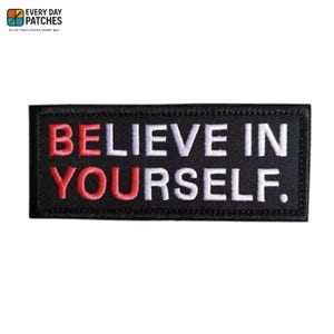 Pode incluir: Patch retangular preto com as palavras "BELIEVE IN YOURSELF" bordadas em fio branco e vermelho. O patch tem uma borda preta e o texto é centralizado. É um acessório motivacional.