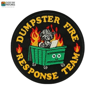 Peut inclure: Un écusson brodé circulaire sur fond noir représentant une benne verte avec des yeux, un raton laveur portant un chapeau et des flammes orange. Le texte jaune DUMPSTER FIRE RESPONSE TEAM forme une bordure circulaire autour du motif.