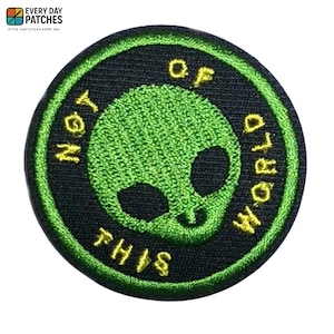 Op de afbeelding: Ronde geborduurde patch met een groene alienkop en de tekst "NOT OF THIS WORLD" in geel. De alienkop staat op een zwarte achtergrond met een groene rand. Een leuk accessoire.