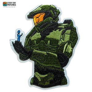 Może przedstawiać: Haftowana naszywka przedstawiająca zielono-czarną opancerzoną postać z pomarańczowym wizjerem, trzymającą małą niebieską figurkę. Naszywka ma białą obwódkę i tekst "EVERY DAY PATCHES" u góry.