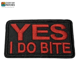May include: Black rectangular patch with red embroidered text reading "YES I DO BITE". The patch has a textured surface and a black border. A fun accessory.
