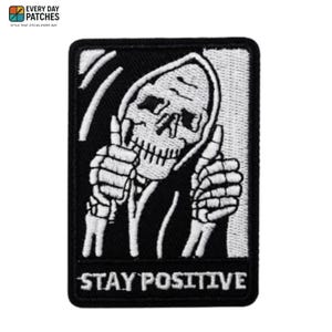Puede incluir: Parche bordado en blanco y negro que representa un esqueleto con una túnica con capucha haciendo un gesto de pulgar hacia arriba. El texto "STAY POSITIVE" se muestra en la parte inferior. El parche tiene forma rectangular con esquinas redondeadas.