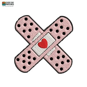 May include: An embroidered patch shaped like two crossed adhesive bandages. The bandages are light pink with a pattern of small black dots and a central white rectangular pad featuring a small, solid red heart stitched in the center.