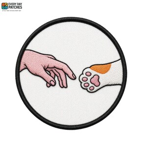May include: A circular embroidered patch featuring a white background with a black border. The design depicts a human hand reaching towards a cat paw, with the human hand in light pink thread and the cat paw in white with pink pads.
