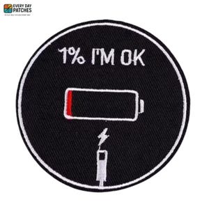 Puede incluir: Parche circular negro con borde blanco. El parche presenta el texto "1% I'M OK" encima de un icono de batería baja con relleno rojo y un símbolo de carga. Un cable de carga blanco en la parte inferior.