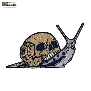 Puede incluir: Parche bordado que representa un caracol con una concha en forma de calavera. La calavera es de color beige con cuencas de ojos y dientes negros. El cuerpo del caracol es gris con detalles en blanco y negro. El parche tiene el texto "EVERY DAY PATCHES".