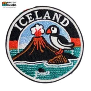 Può includere: Toppa ricamata con la scritta "ICELAND" in bianco, arcuata sopra una scena di un vulcano in eruzione con lava rossa e un pulcinella di mare. La toppa ha un bordo nero con accenti rossi e verde acqua.