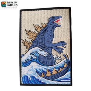 Pode incluir: Patch bordado com um design azul de Godzilla emergindo de uma onda. O patch tem uma borda preta e o texto "EVERY DAY PATCHES" no canto superior esquerdo. Godzilla tem uma boca vermelha e barbatanas dorsais castanhas.