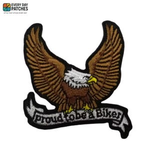 Pode incluir: Patch bordado com uma águia marrom e bege com as asas abertas. A águia tem cabeça branca e bico amarelo. Uma faixa abaixo diz "Proud to be a Biker" em letras brancas. O patch tem uma borda preta.