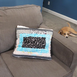 May include: A decorative square pillow with a patchwork design rests on a gray sofa. The pillow features a central black and white patterned square, surrounded by light blue and gray fabric strips. A dog is lying on the floor in the background.