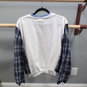 May include: White sweatshirt with blue plaid sleeves and a light blue collar. The back of the shirt is visible, hanging on a black hanger against a neutral wall.