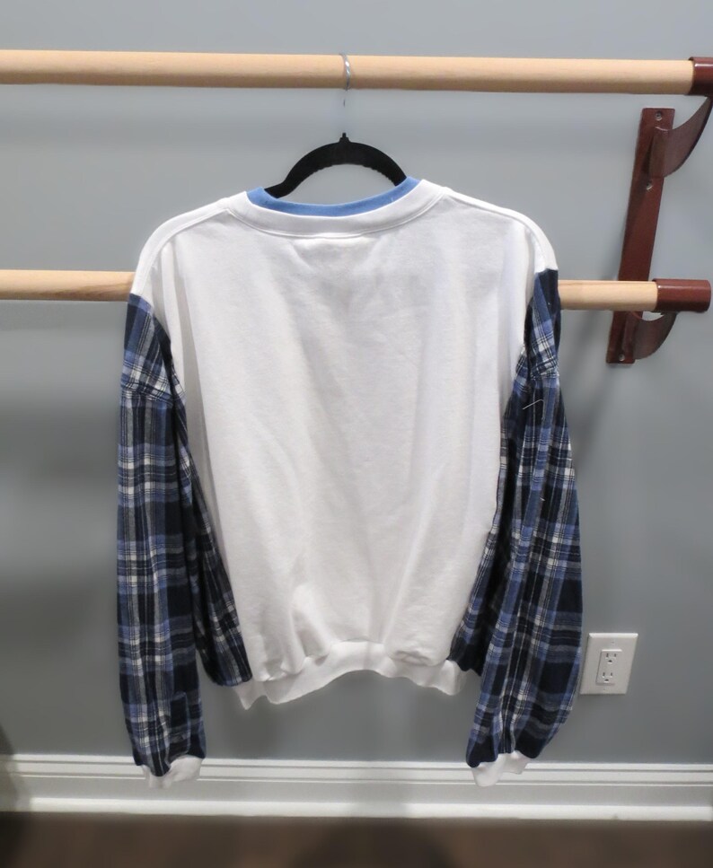 May include: White sweatshirt with blue plaid sleeves and a light blue collar. The back of the shirt is visible, hanging on a black hanger against a neutral wall.