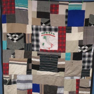 May include: A patchwork quilt made from various fabrics, including plaid, striped, and solid-colored materials. The quilt features a central square with an Italian flag design. The quilt is bordered with a black fabric and hung with clothespins.