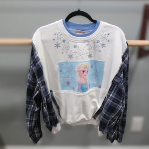 May include: White sweatshirt with blue plaid sleeves and a light blue collar. The front features a rectangular image of a cartoon character with blonde hair and a blue dress, surrounded by snowflakes.