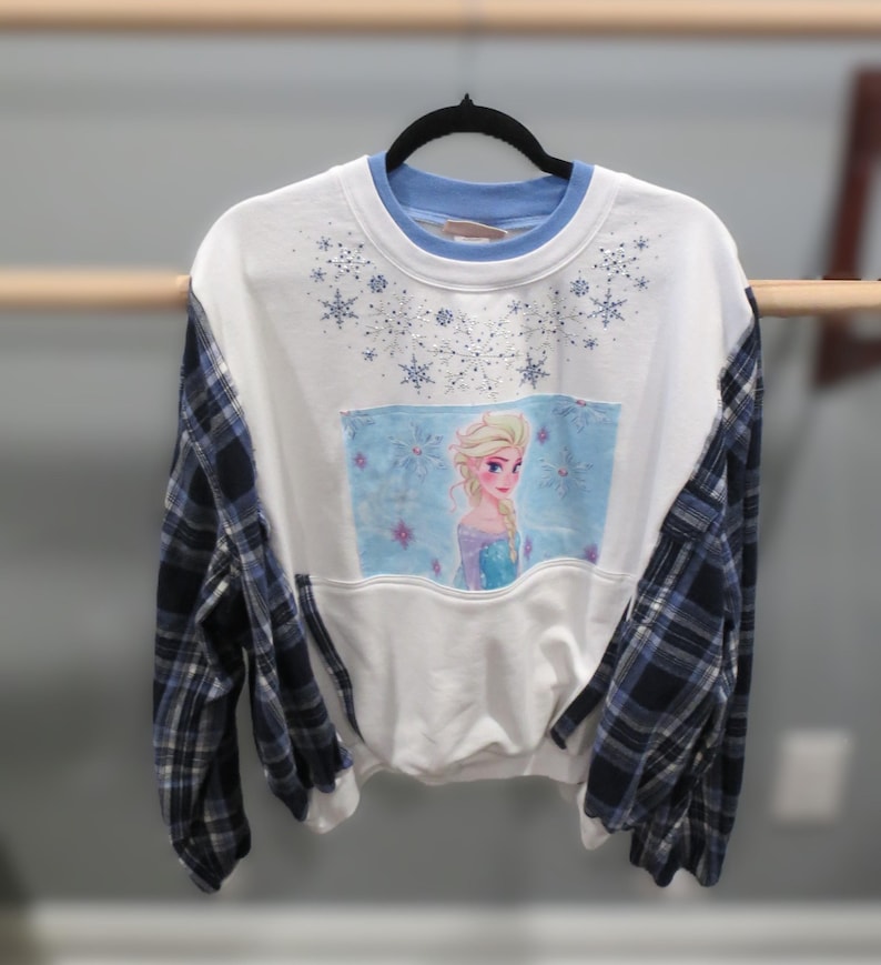 May include: White sweatshirt with blue plaid sleeves and a light blue collar. The front features a rectangular image of a cartoon character with blonde hair and a blue dress, surrounded by snowflakes.