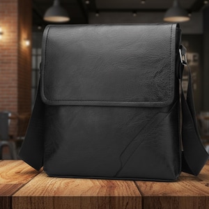 May include: A black messenger bag with a flap closure and adjustable shoulder strap. The bag is made of textured material and sits on a wooden surface. The bag has a rectangular shape and a diagonal design element.