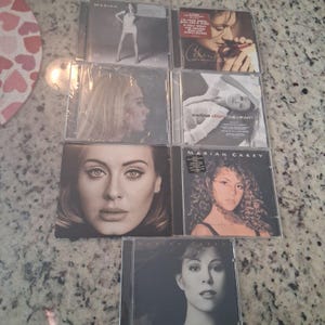 May include: A collection of music CDs featuring album covers with portraits of female singers. The CDs are in clear plastic cases, with images of Mariah Carey, Adele, and Celine Dion. The album covers are in various shades of black, white, and sepia.