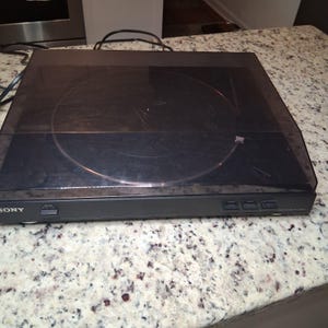 Może przedstawiać: Czarny gramofon Sony z przezroczystą, otwieraną pokrywą przeciwkurzową. Gramofon posiada okrągły talerz i przyciski sterowania na przednim panelu. Urządzenie stoi na nakrapianym blacie.