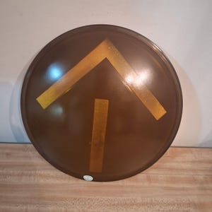 May include: A round, brown shield with a gold arrow design. The shield has a glossy finish and is set against a light wood surface. The arrow is composed of a horizontal bar and a vertical bar.