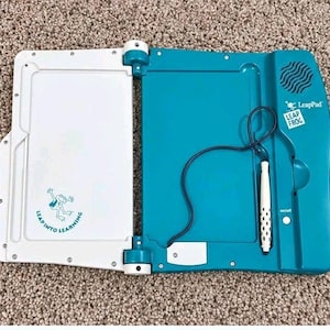 Puede incluir: Una tableta de aprendizaje LeapPad en color verde azulado y blanco con un lápiz óptico blanco unido por un cable negro. El dispositivo cuenta con una pantalla blanca con el logotipo de LeapFrog y el texto Leap Into Learning, y un panel de control.