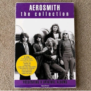 May include: A rectangular Aerosmith The Collection 3 CD box set featuring a black and white photograph of the band members. A yellow circular sticker on the front lists 21 original tracks including Dream On, Mama Kin, and Walk This Way.