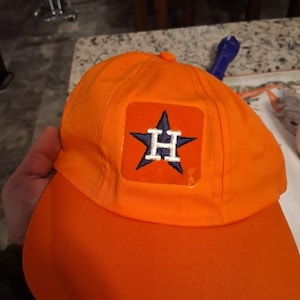 May include: Bright orange baseball cap featuring a square patch with a navy blue star and a white letter "H". The cap has a curved brim and a rounded top. This cap is a sports fan accessory.