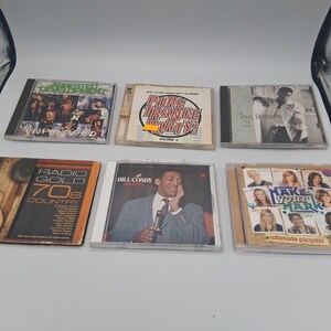 May include: A collection of six CDs, including albums by Arrested Development, Jon Secada, and Bill Cosby. The CDs are in their original jewel cases, with album titles and artist names visible. The image is taken from a high angle.