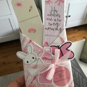 Puede incluir: Dos marcapáginas rectangulares de papel dentro de un sobre con estampado rosa y blanco. Un marcapáginas presenta una pequeña flor, una seta y el texto BLESSED BEYOND MEASURE. El otro presenta lazos rosas y el texto His mercies are new every morning.