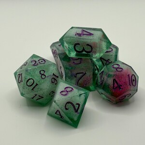 Op de afbeelding: Een set van zeven groene polyhedrale dobbelstenen met paarse cijfers. De dobbelstenen hebben een glitterachtig uiterlijk en zijn ontworpen voor rollenspellen. De dobbelstenen liggen op een witte ondergrond.
