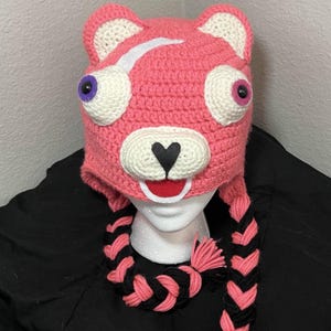 Op de afbeelding: Een roze gehaakte berenmuts met witte accenten, grote cartoonogen en zwart-roze gevlochten oorflappen. De muts heeft een zwarte hartneus en een rode mond. De muts is ontworpen om op het hoofd te worden gedragen.