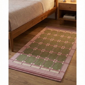 May include: A rectangular rug with a green and pink plaid pattern. The rug has a pink border and is placed on a wooden floor. The rug is near a wooden bed frame and a bedside table.