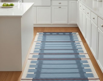 May include: A rectangular blue and white patterned rug with a geometric design. The rug features horizontal stripes in varying shades of blue, accented with white and a dark red border. It is placed on a hardwood floor in a kitchen setting.