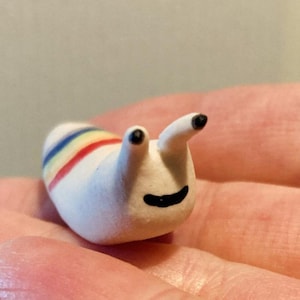 May include: A small, white ceramic snail figurine with a rainbow stripe design on its back. The snail has two black eyes and a smiling black mouth. The figurine is held in a person's hand.