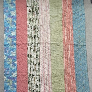 May include: A rectangular quilt featuring vertical panels of varying fabric patterns and colors. The design includes blue floral, solid dusty rose, cream with dark text, thin red stripes on white, sage green gingham, and red geometric print fabric.