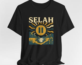 Puede incluir: Camiseta negra con un diseño inspirado en la naturaleza. El gráfico muestra un sol amarillo con un símbolo de pausa, montañas y agua. La palabra "SELAH" está impresa encima del sol.