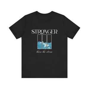 Könnte beinhalten: Schwarzes T-Shirt mit dem Wort „STRONGER“ über einer Grafik aus drei vertikalen Rechtecken mit blauen Wellen und weißen Details. Darunter steht der Text „than the storm“.