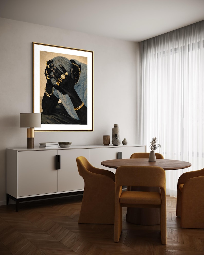May include: A framed art print depicts a person with their face obscured by their hands. The person is adorned with gold jewelry, including rings and bracelets. The artwork is displayed in a room with a white cabinet, a round wooden table, and mustard-colored chairs.