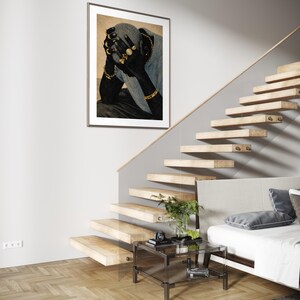 May include: A framed art print featuring a person with dark skin, adorned with gold jewelry, covering their face. The artwork hangs on a white wall near a wooden staircase and a glass side table.