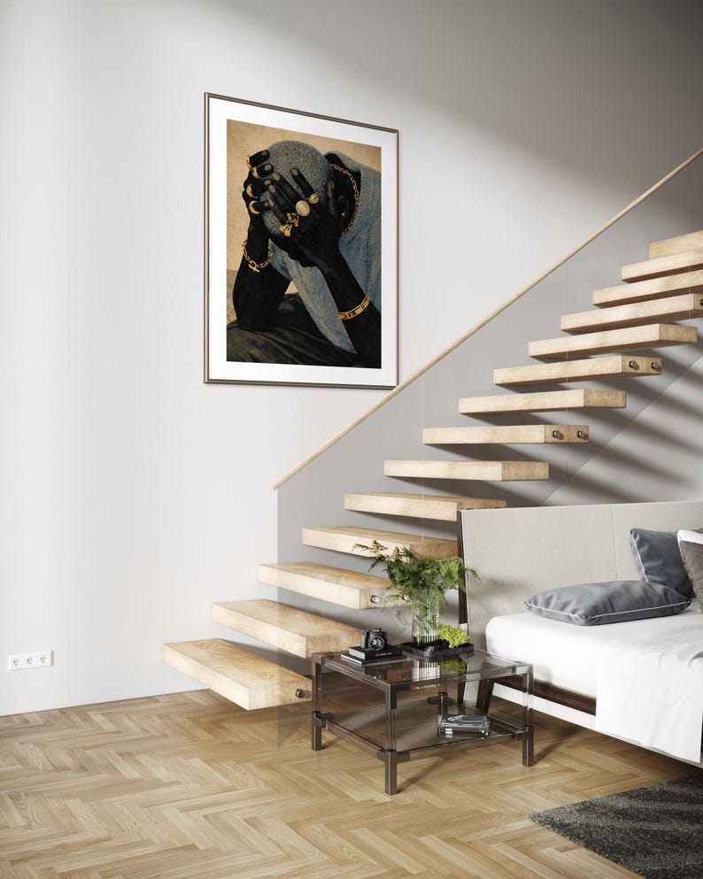 May include: A framed art print featuring a person with dark skin, adorned with gold jewelry, covering their face. The artwork hangs on a white wall near a wooden staircase and a glass side table.