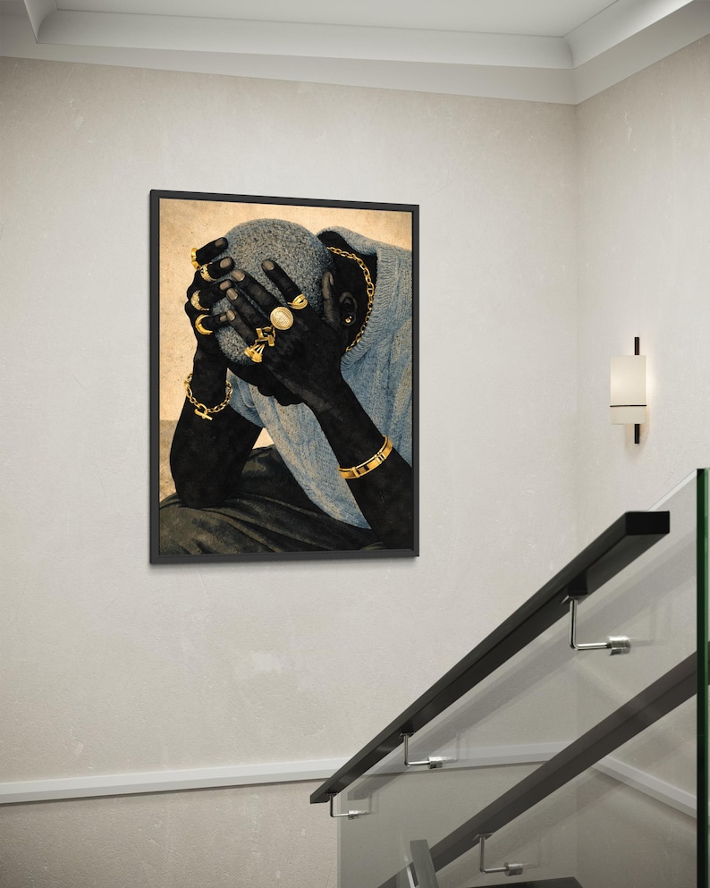 May include: Framed art print featuring a person with dark skin, wearing gold jewelry, including rings, bracelets, and a chain. The person is wearing a blue sweater and has their hands covering their face. The artwork is displayed on a white wall.