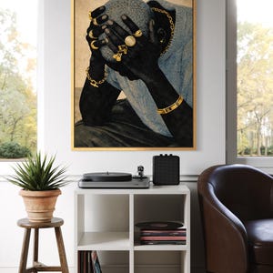 May include: A framed art print depicts a person with dark skin, wearing gold jewelry, and a light blue sweater. The artwork is displayed above a white cube shelf with a record player, speaker, and potted plant.
