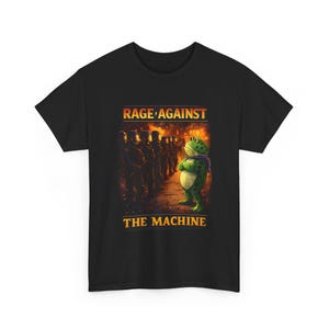 Puede incluir: Camiseta negra con una rana de dibujos animados con una bufanda morada, de pie frente a una fila de soldados. El texto "RAGE AGAINST THE MACHINE" está impreso encima y debajo de la ilustración de la rana en naranja.