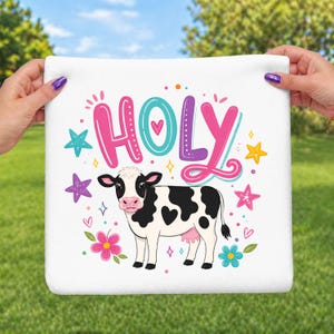 May include: White square towel with a cartoon cow illustration. The cow is black and white with heart-shaped spots. The word "HOLY" is written in colorful bubble letters above the cow, surrounded by stars, hearts, and flowers.