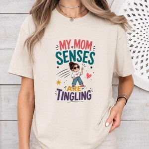 Könnte beinhalten: Beiges T-Shirt mit dem Text "MY MOM SENSES ARE TINGLING" in Rosa, Grün und Lila. Die Grafik zeigt ein Cartoon-Mädchen, Sterne und Herzen. Ein lässiges T-Shirt für den Alltag.