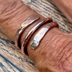 May include: A copper-coloured bypass ring featuring a triple-wrap design. The metal band displays a hammered, textured finish on the two outer ends, while the central section remains smooth and polished, creating a distinct contrast in surface detail.