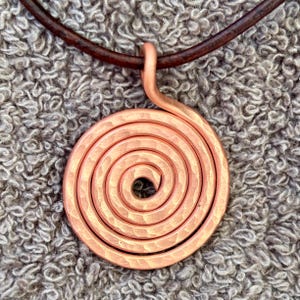 May include: A copper-coloured metal pendant shaped as a flat spiral with a hammered, textured surface. The pendant is suspended from a dark brown, smooth, rounded cord that forms a circular loop at the top of the spiral design.