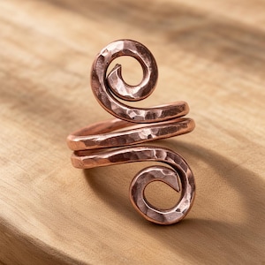 May include: A copper ring featuring a bypass design with two spiraling ends. The metal surface has a distinct hammered texture, creating a series of small, reflective indentations across the entire length of the coiled band structure.