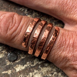 May include: A copper ring featuring a wrap-around design with four distinct bands. The metal surface has a hammered, textured finish that reflects light, creating a metallic sheen across the curved, continuous structure of the jewellery piece.