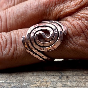 May include: A copper-toned ring featuring a prominent spiral design. The metal surface displays a distinct hammered texture throughout, creating a series of small indentations that catch the light across the curved, overlapping band structure.