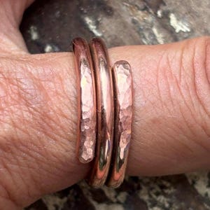 May include: A copper ring featuring a wrap-around design with three distinct bands. The metal surface has a hammered, textured finish that catches the light, creating a reflective appearance across the reddish-brown metallic material of the ring.